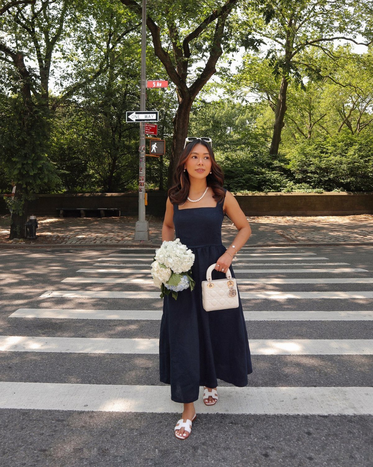 Woman in a navy blue summer dress and white sandals walking across a city street.