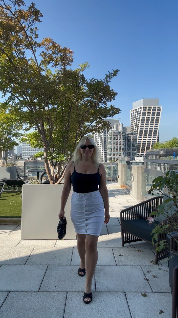 A blonde woman in a black tank top and white skirt walking on a sunny rooftop.