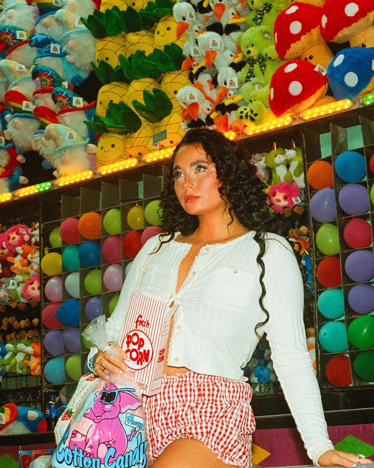 A young woman in checkered shorts holding cotton candy at a colorful carnival game stall.