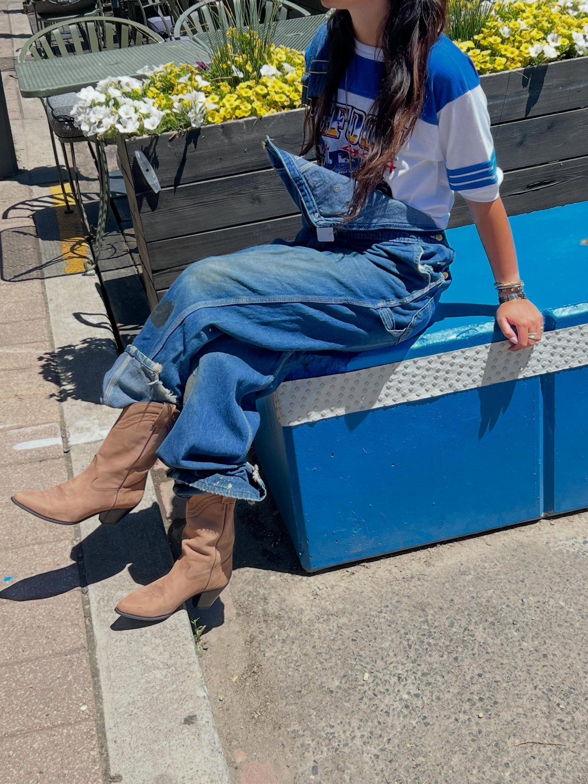 Person sitting on a blue ledge wearing denim overalls, a graphic tee, and tan boots.