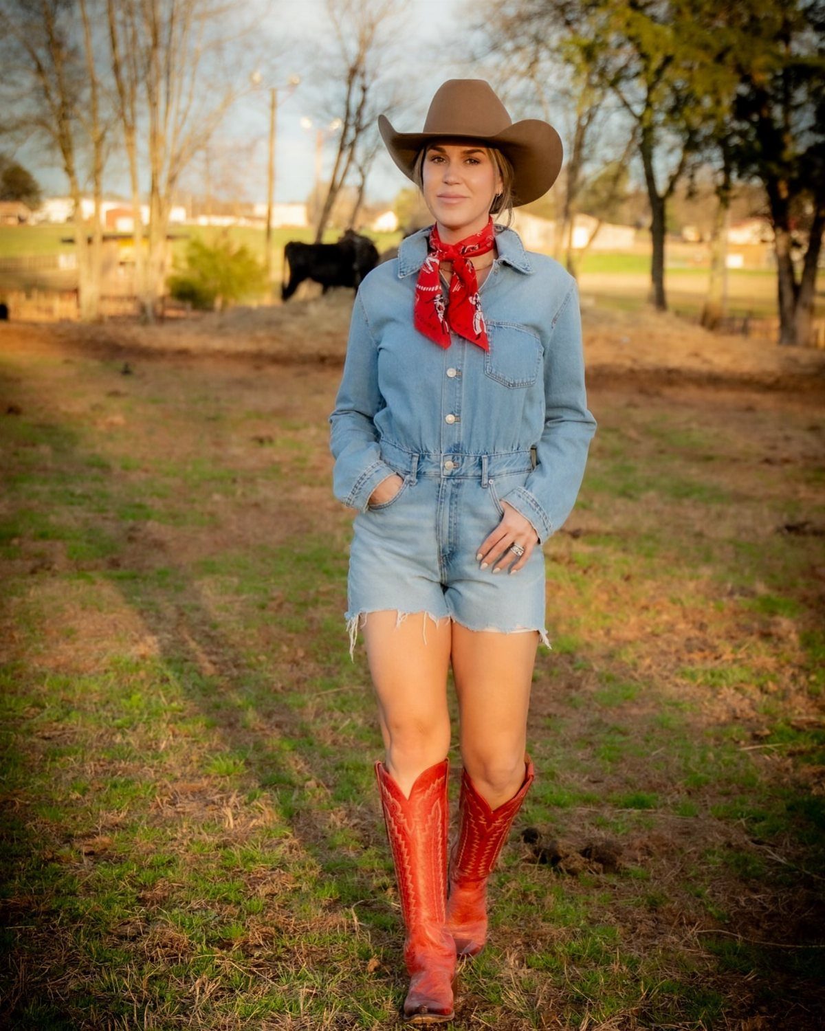 Woman in a denim romper, red bandana, cowboy hat, and red boots standing in a field.