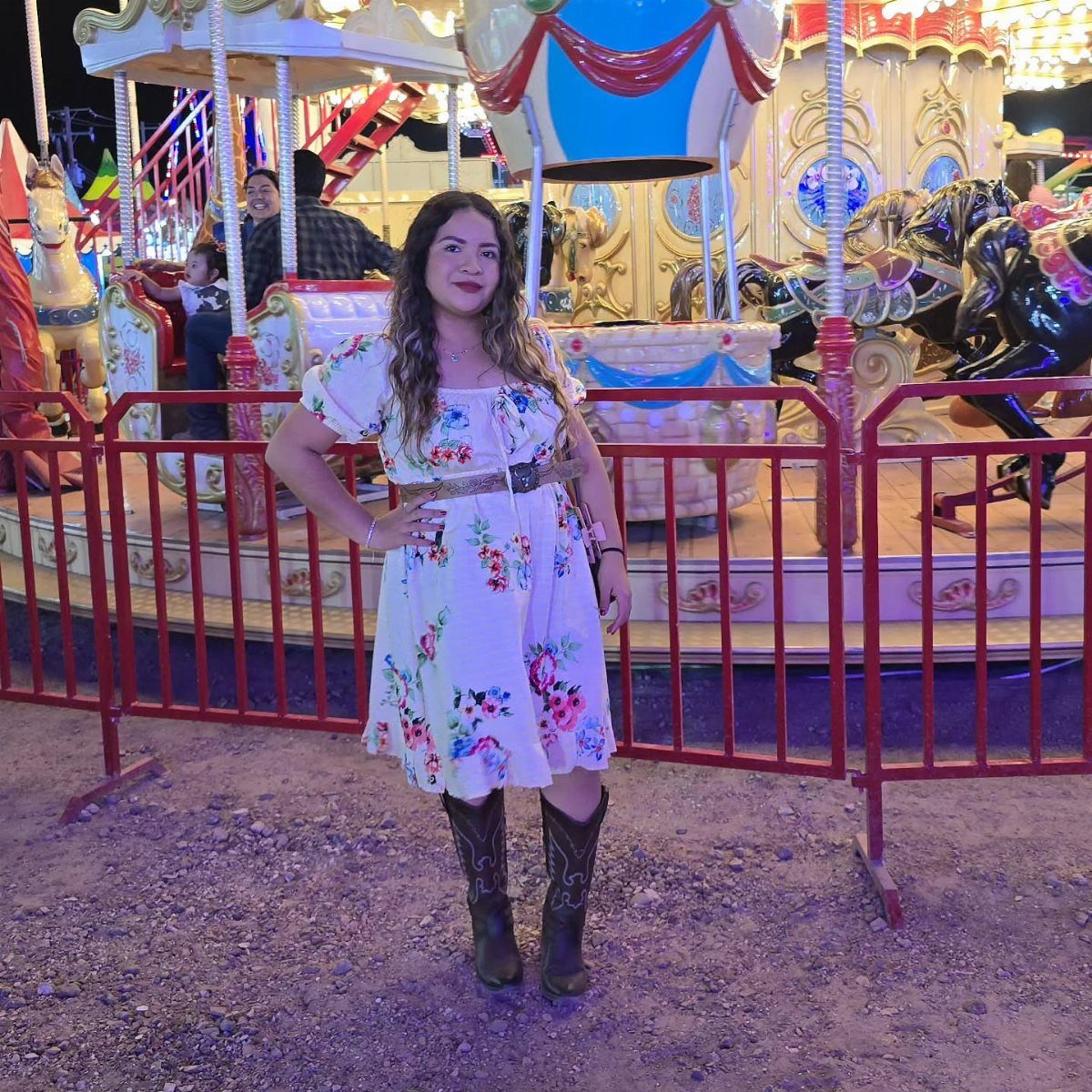 Woman in a white floral midi dress and cowgirl boots standing before a lit carousel.