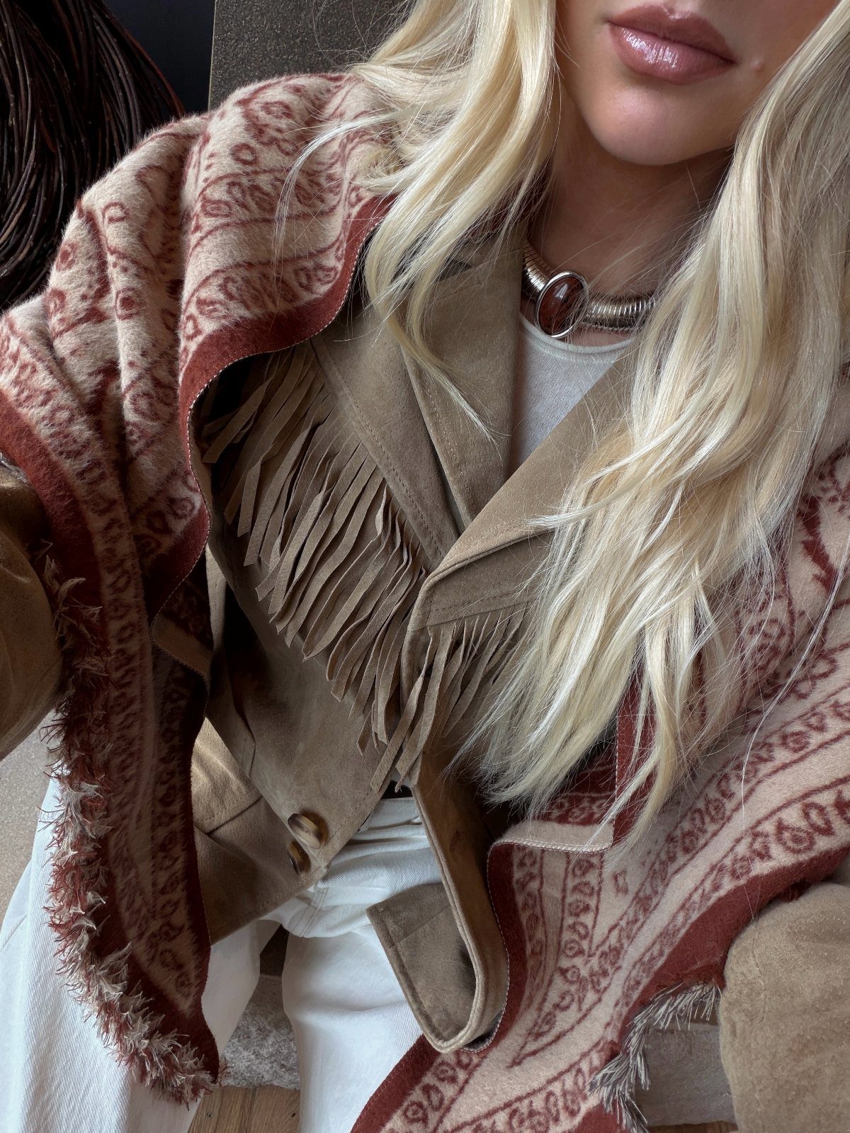 Blonde woman wearing a fringed suede jacket and a patterned red and beige shawl.