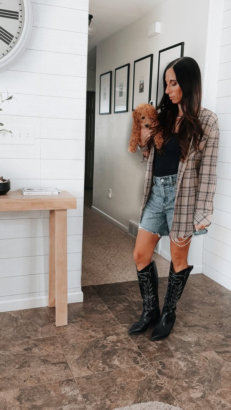 Woman in a plaid shirt, denim shorts, and black cowboy boots holding a small dog.