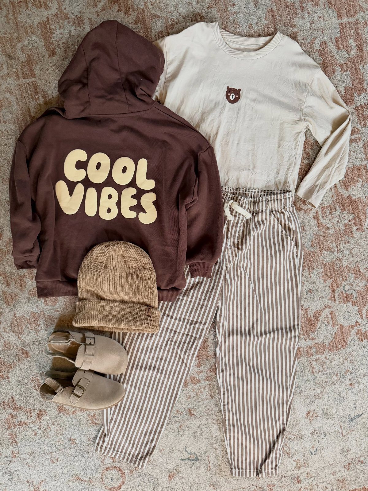 Children's clothing flat lay featuring a brown hoodie, cream shirt, striped pants, beanie, and clogs.