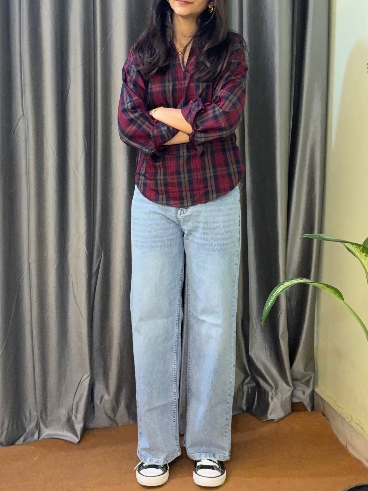 A person wearing a red plaid shirt and wide-leg blue jeans standing before grey curtains.