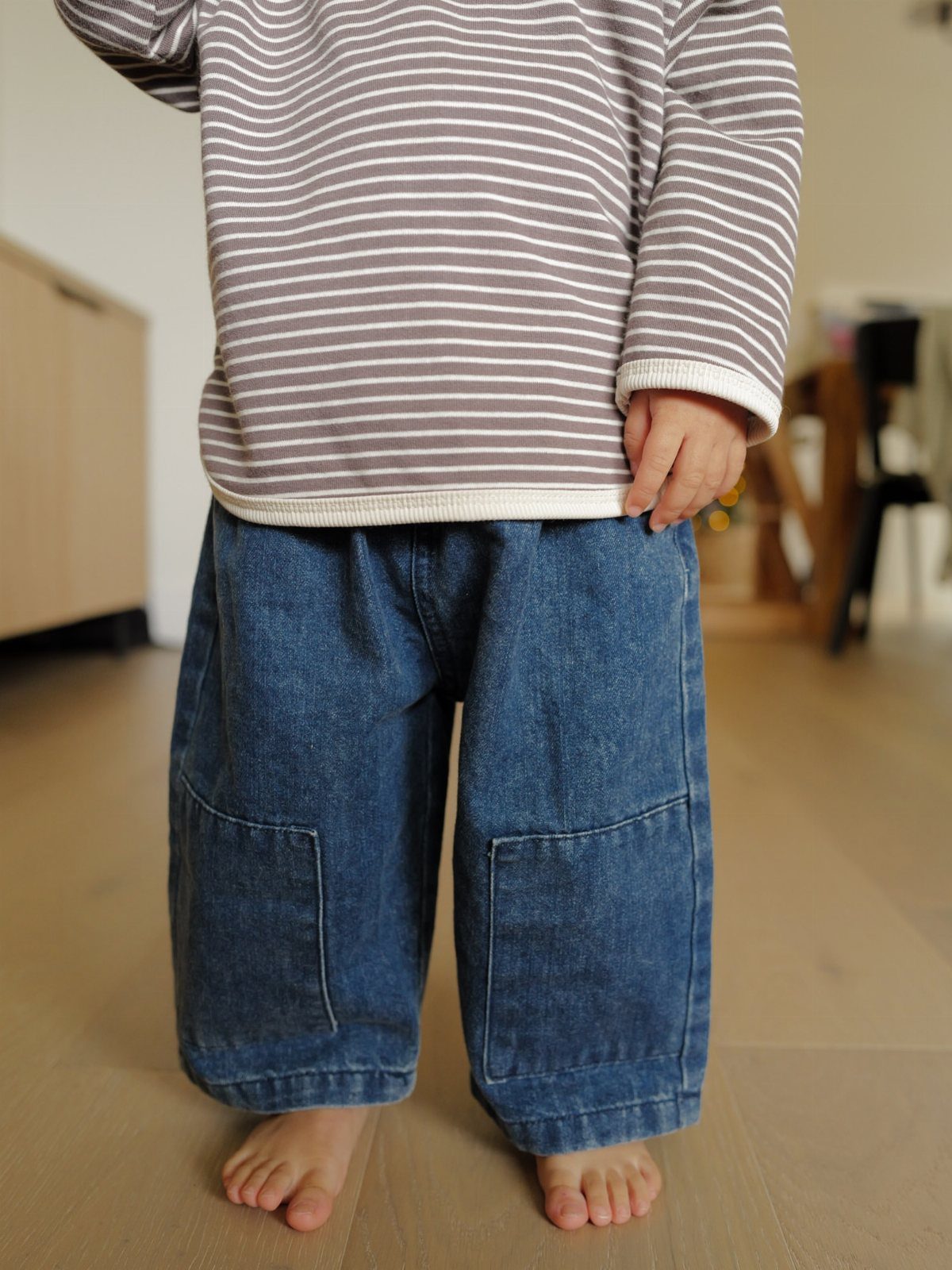 Child wearing a striped long-sleeved shirt and wide blue denim trousers standing barefoot.