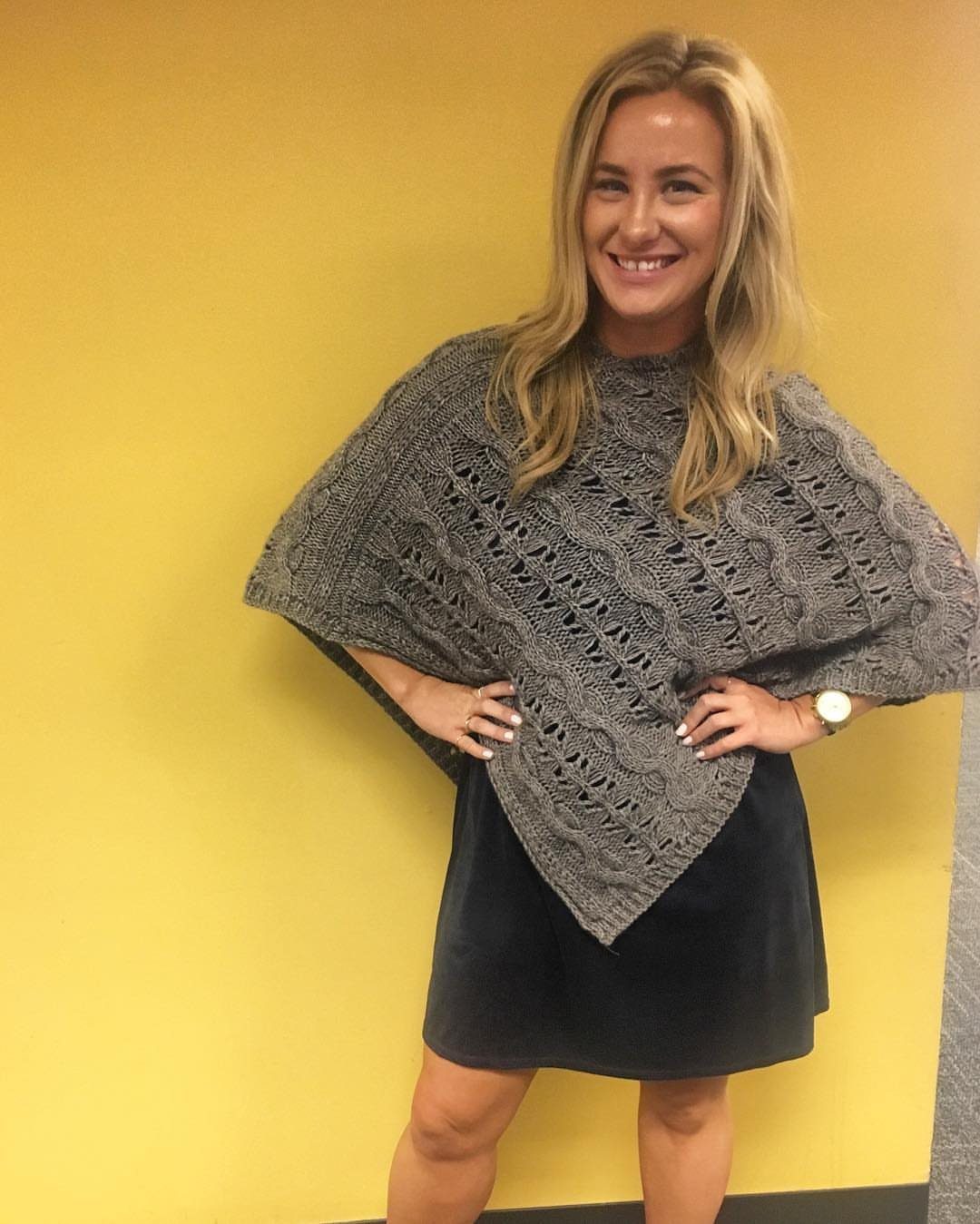 A smiling blonde woman wearing a grey knit poncho and a black skirt against a yellow wall.