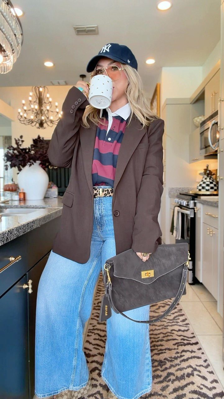 Woman in a brown oversized blazer, striped shirt, and wide-leg jeans holding a white mug.