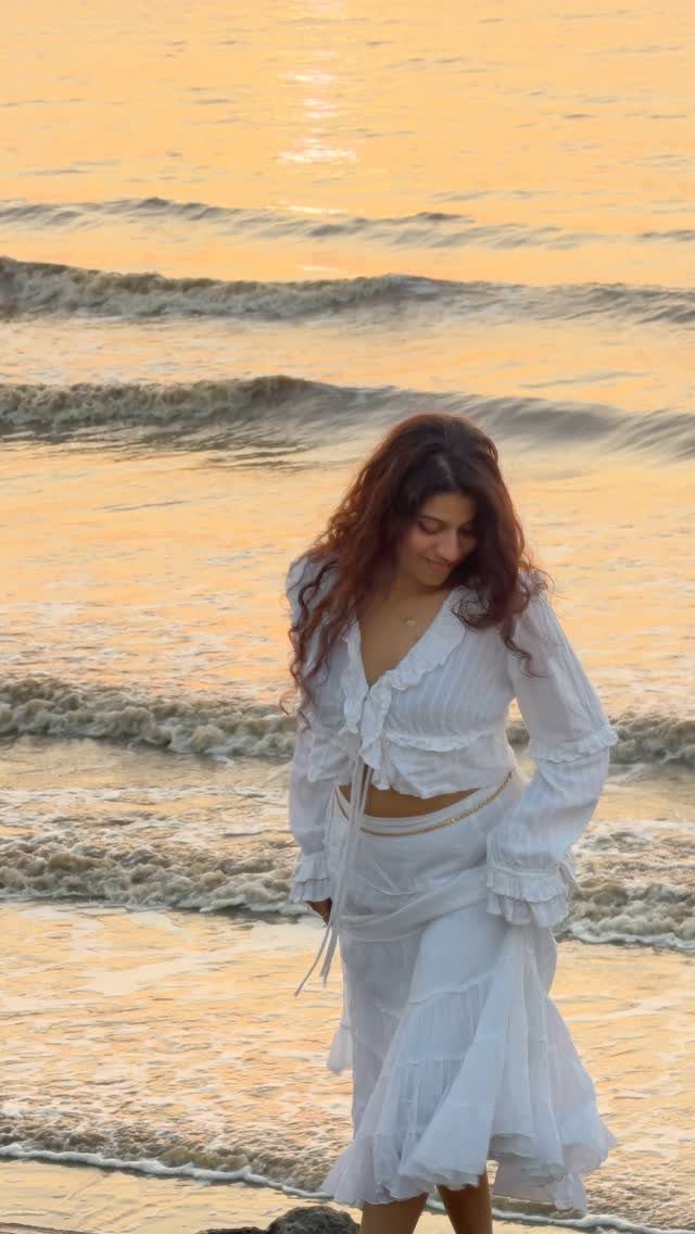 Woman in white ruffled crop top and maxi skirt walking on a sunset beach.