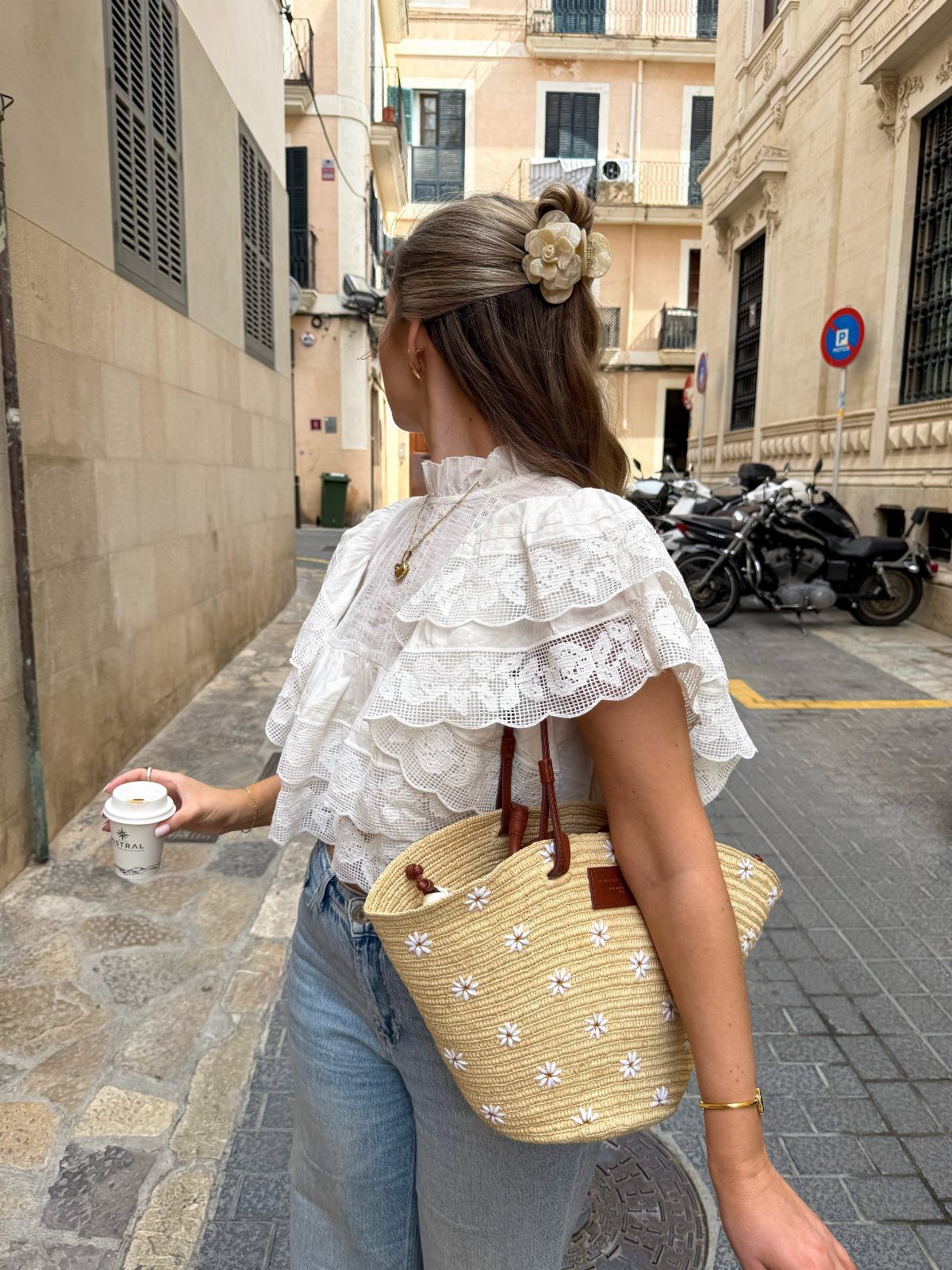 Woman in a white lace blouse and denim jeans walking through a narrow street with a straw bag.