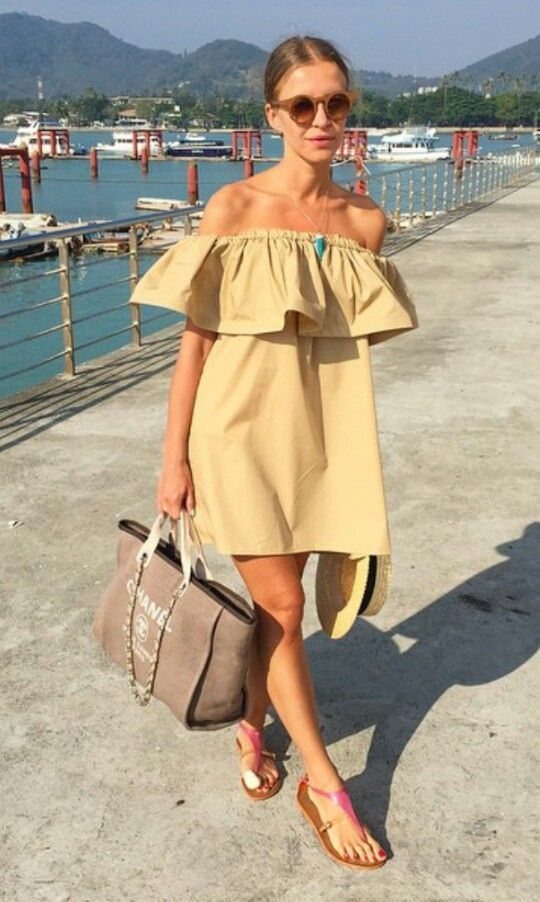 A woman in a tan off-the-shoulder ruffled mini dress walking along a sunny waterfront pier.