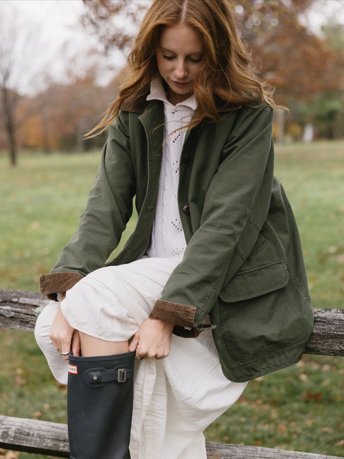 A woman wearing an olive green barn coat and white cable knit sweater sitting on a fence.