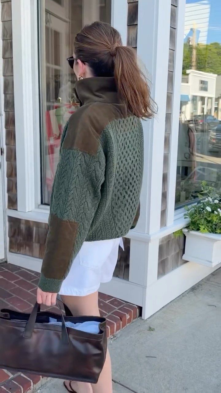 Woman wearing an olive green cable knit sweater with brown patches and white shorts.