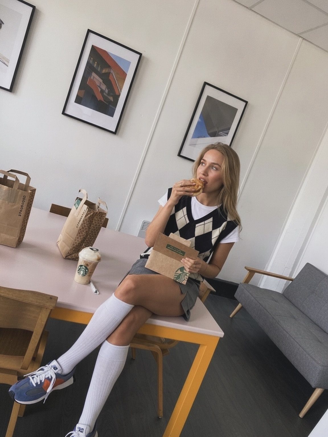 A woman wearing an argyle sweater vest, grey skirt, and white knee-high socks sitting at a table.