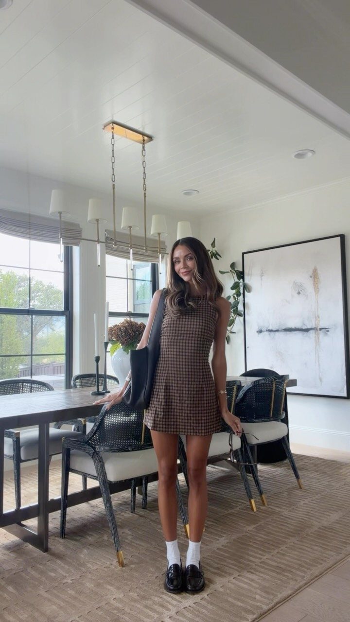 A woman posing in a brown plaid dress, white socks, and black loafers indoors.