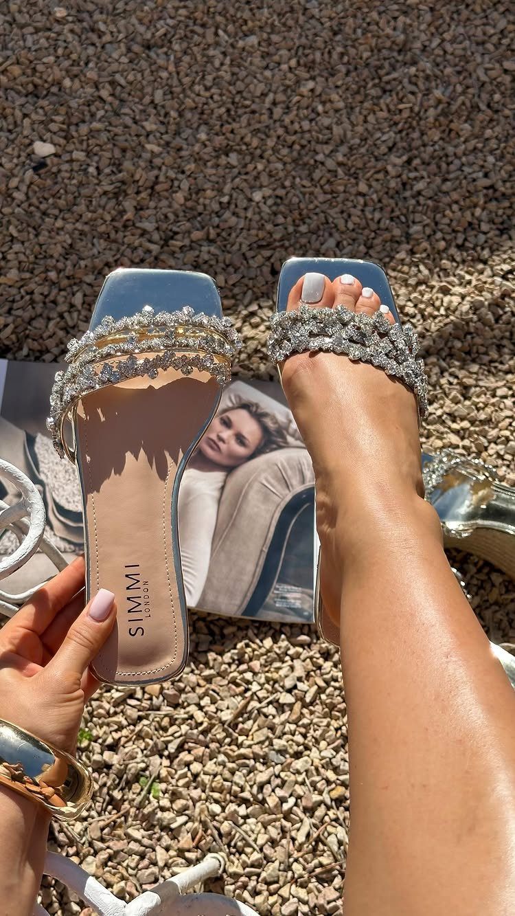 Silver jeweled slide sandals with sparkling rhinestone straps worn on feet over gravel.