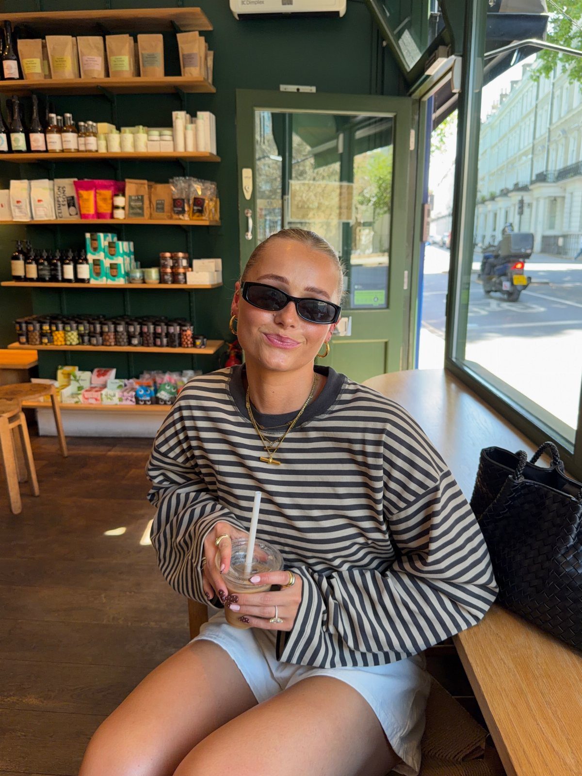 A smiling woman in sunglasses and a striped shirt sitting in a cafe holding a drink.