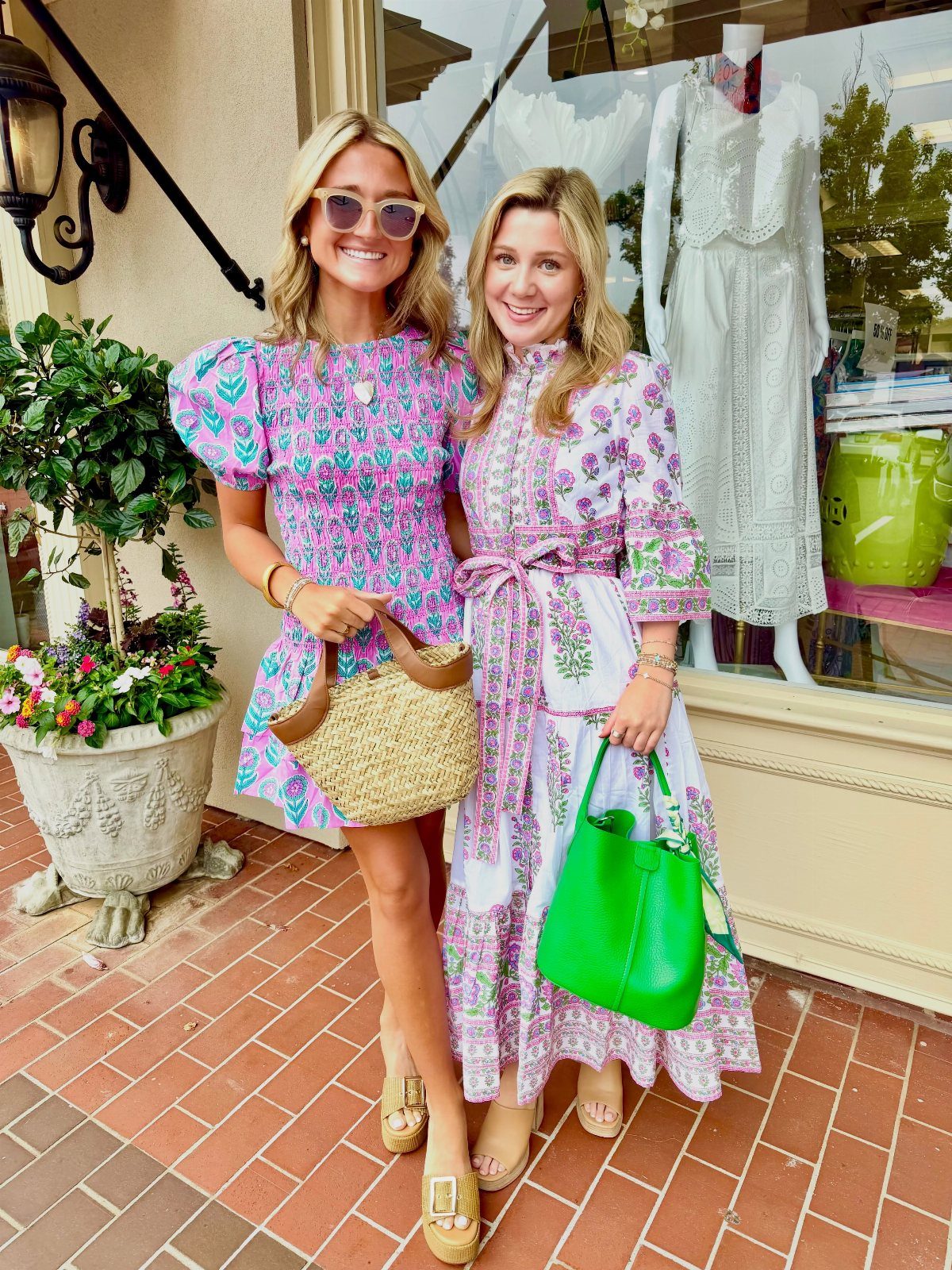 Two women posing in colorful patterned summer dresses and carrying handbags outdoors.