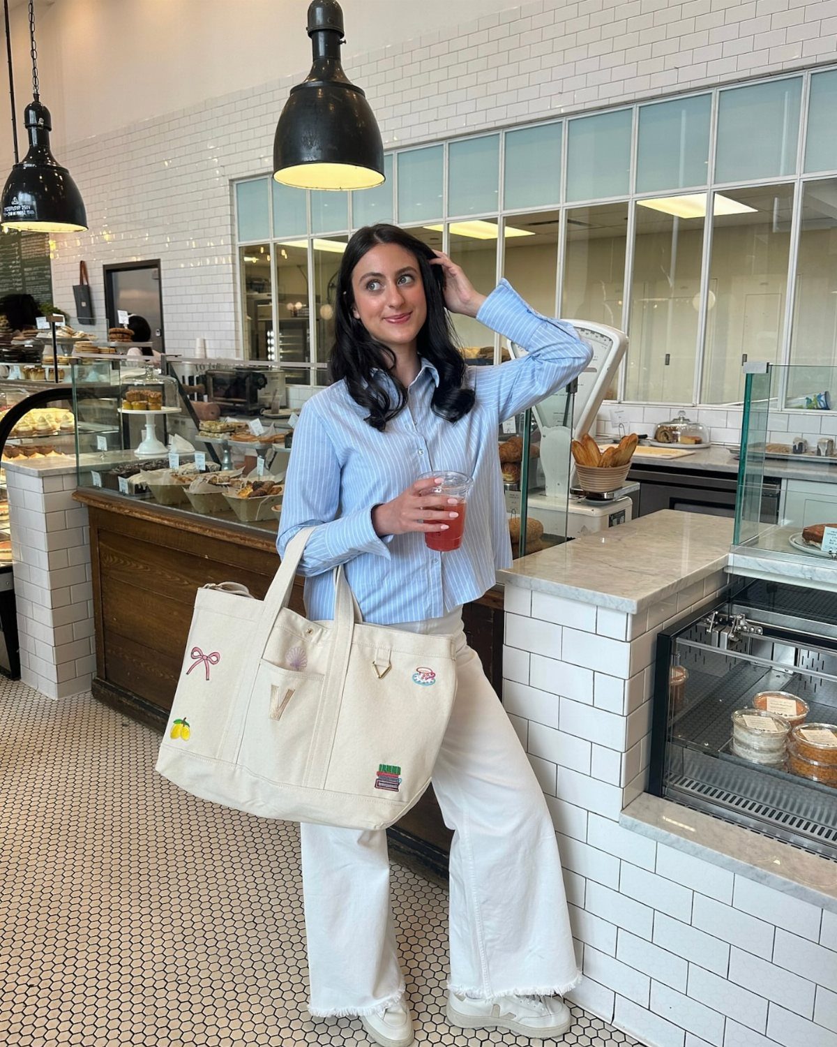 A woman in a blue striped shirt and white pants holding a drink and tote bag.