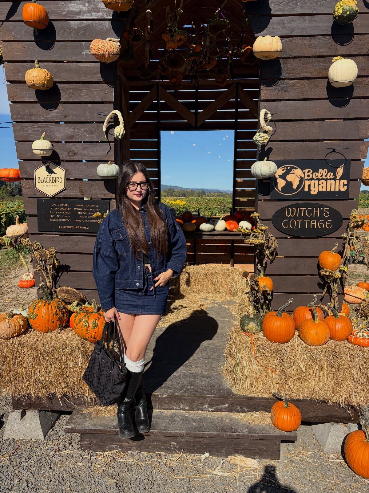 Woman in a denim jacket and skirt standing amidst pumpkins and gourds.