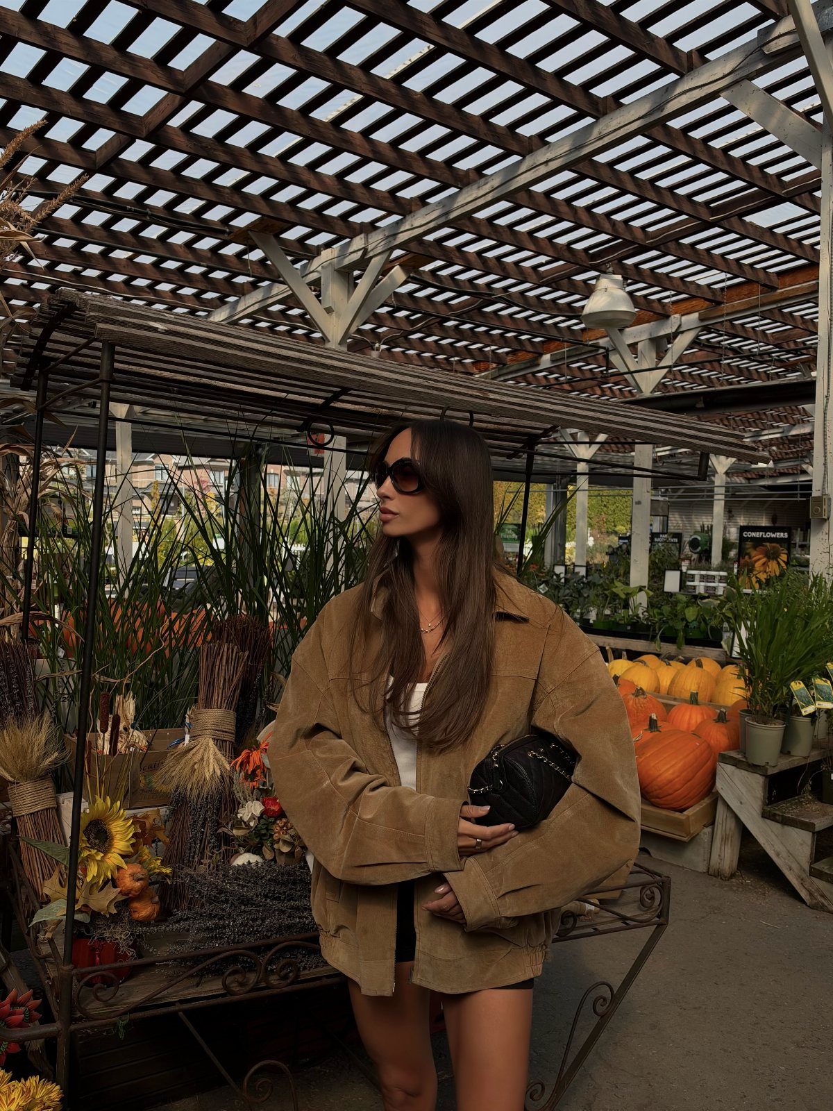 A woman wearing a brown faux suede jacket and sunglasses standing near pumpkins.
