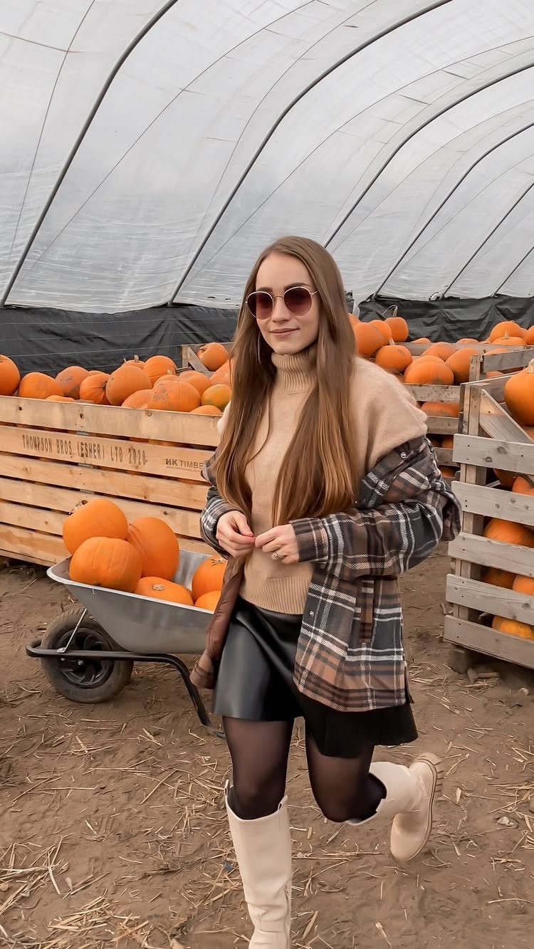 A woman wearing cream knee-high boots posing in a greenhouse filled with pumpkins.