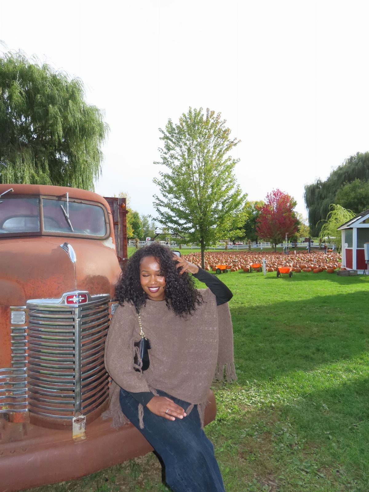A smiling woman wearing a brown knit poncho and denim skirt sitting on a vintage truck.