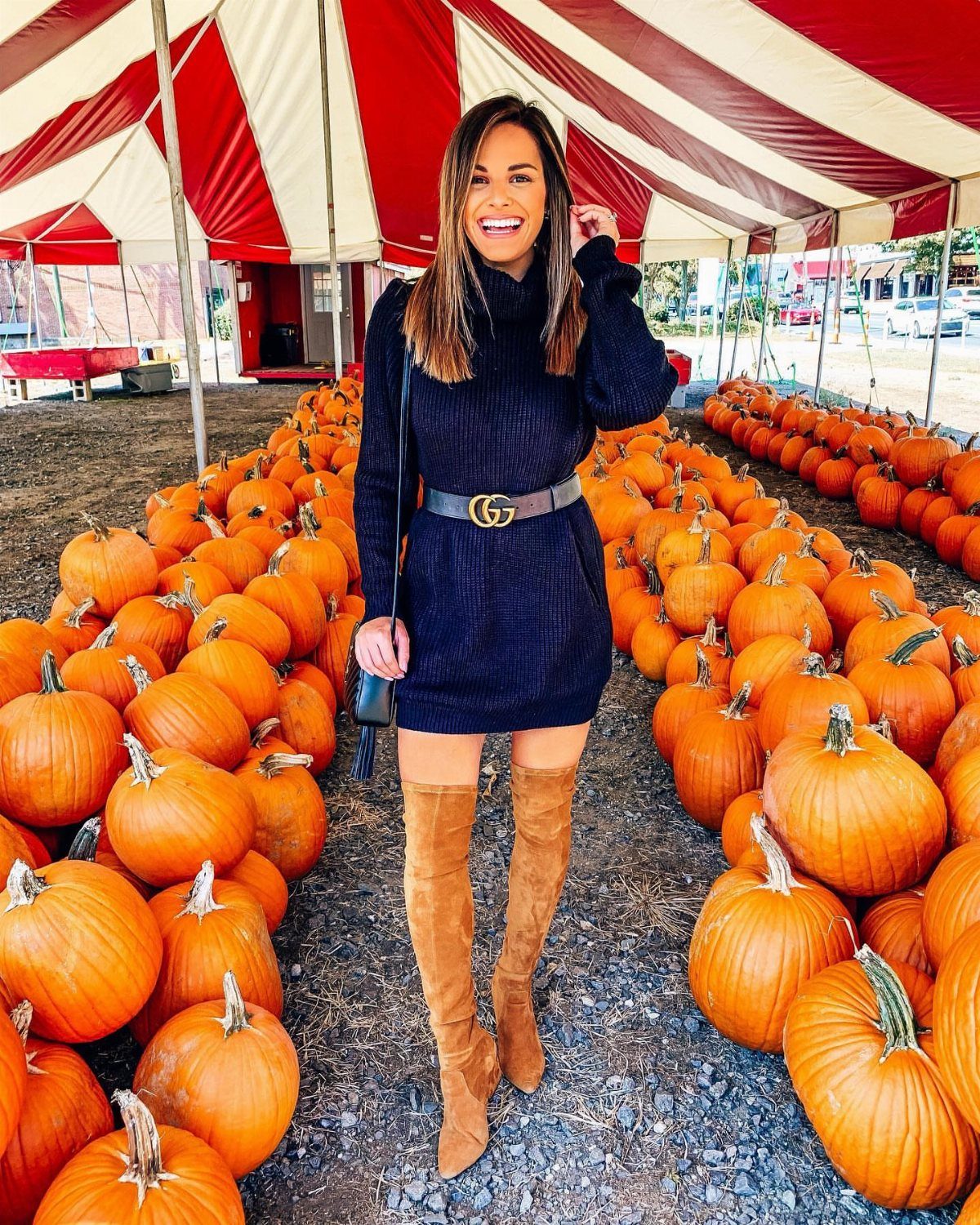 A woman in a black sweater dress and tan boots posing in a pumpkin patch.