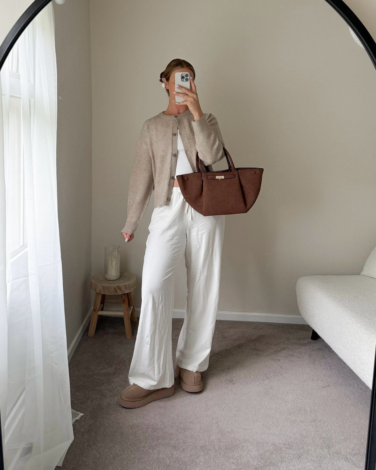 A person posing in a mirror wearing a beige cardigan, white trousers, and carrying a brown handbag.