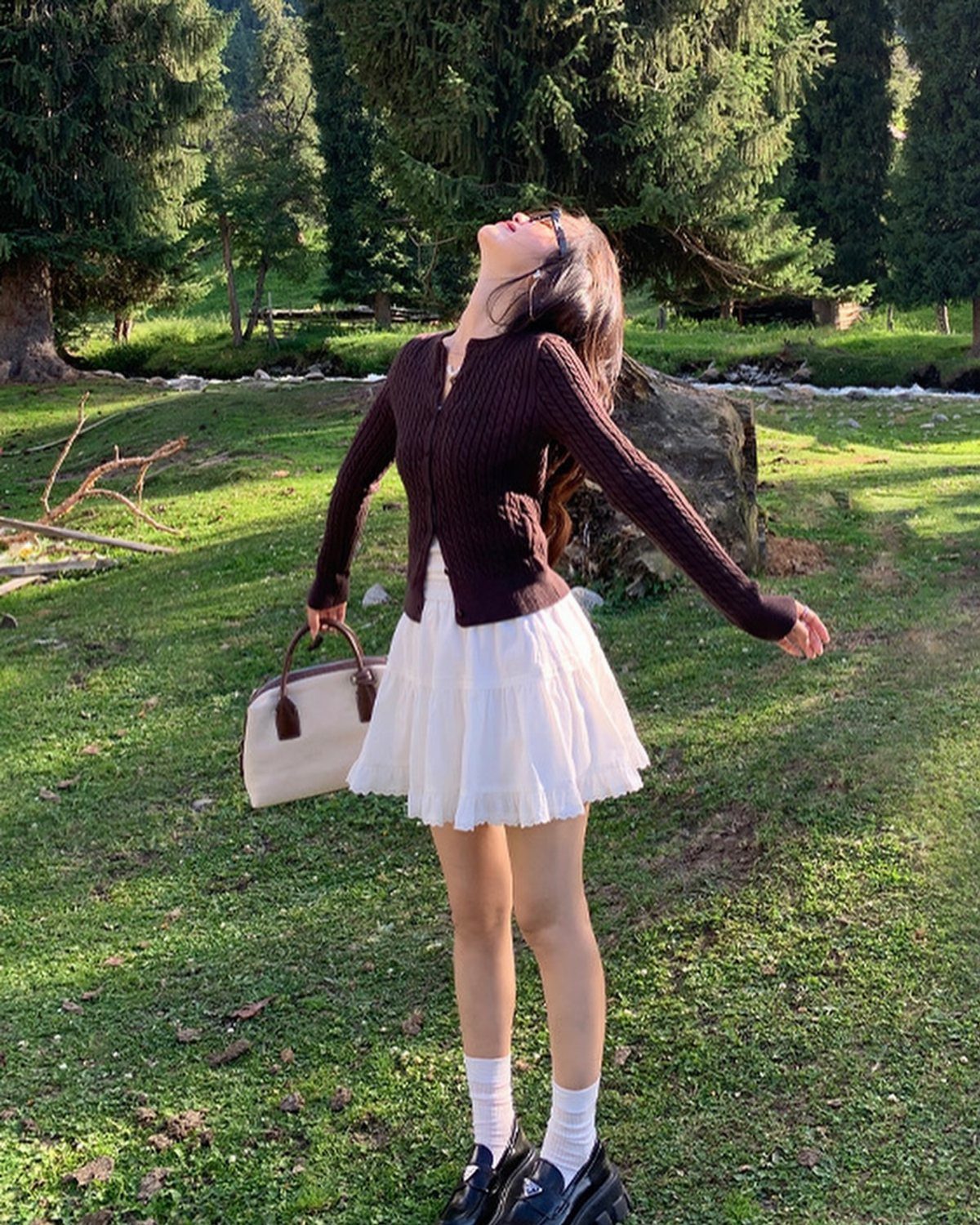 Woman in a brown cardigan and white pleated skirt standing in a sunny grassy field.