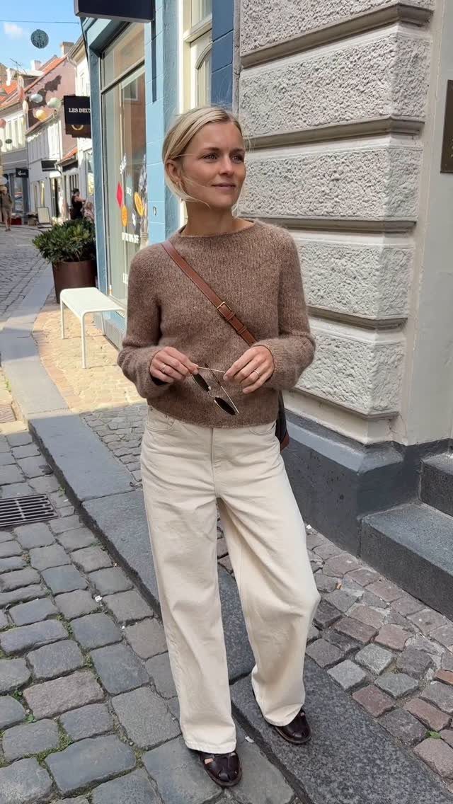 A woman wearing a brown knit sweater and cream trousers standing on a cobblestone street.
