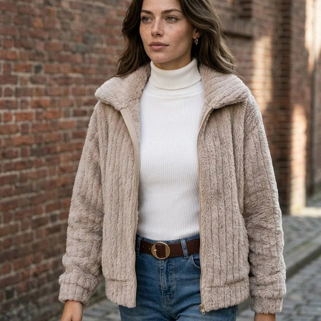 A woman wearing a beige textured jacket, white turtleneck, and blue jeans standing by a brick wall.