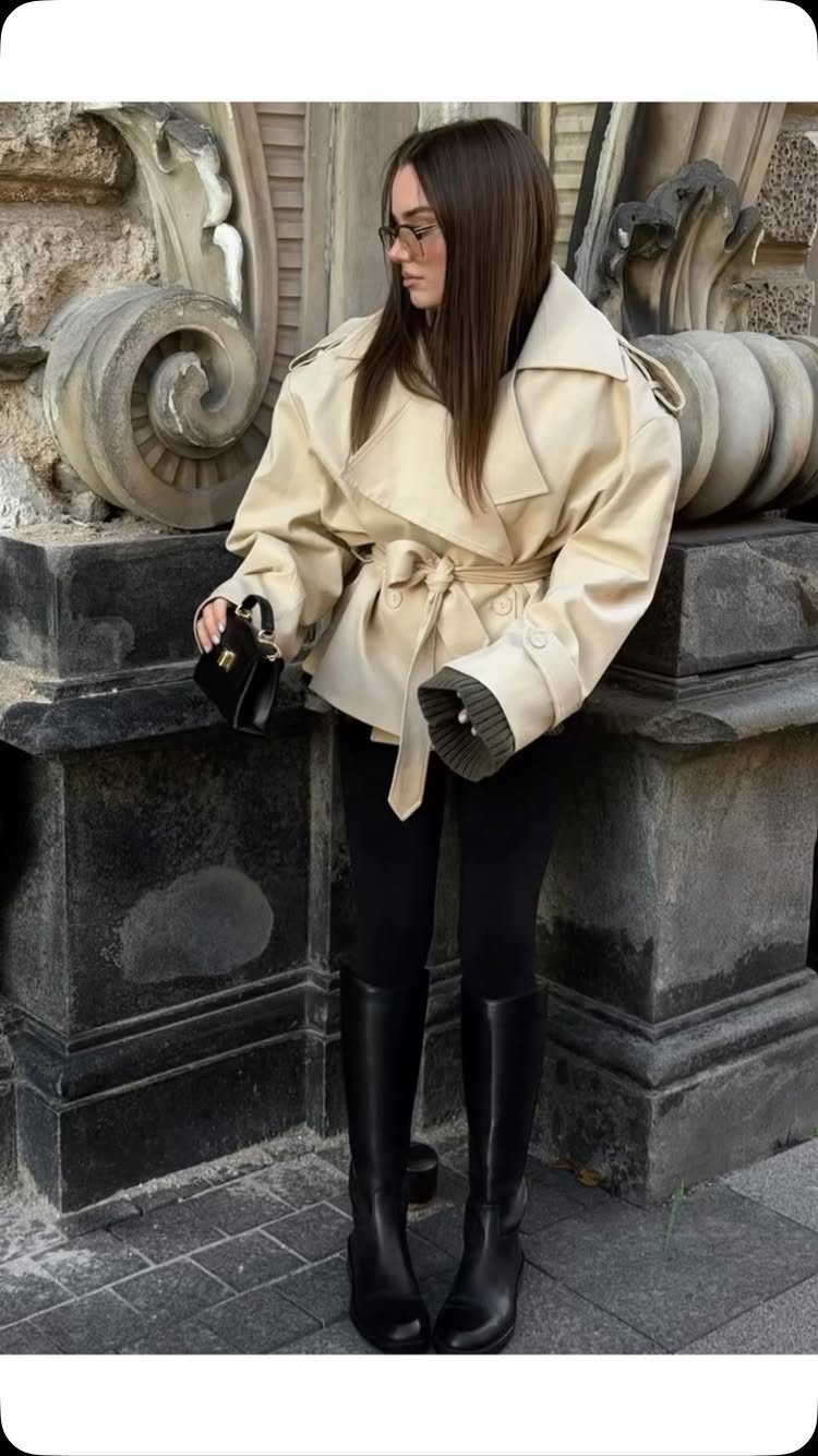 A woman wearing a cream trench coat, black leggings, and black boots standing by a stone building.
