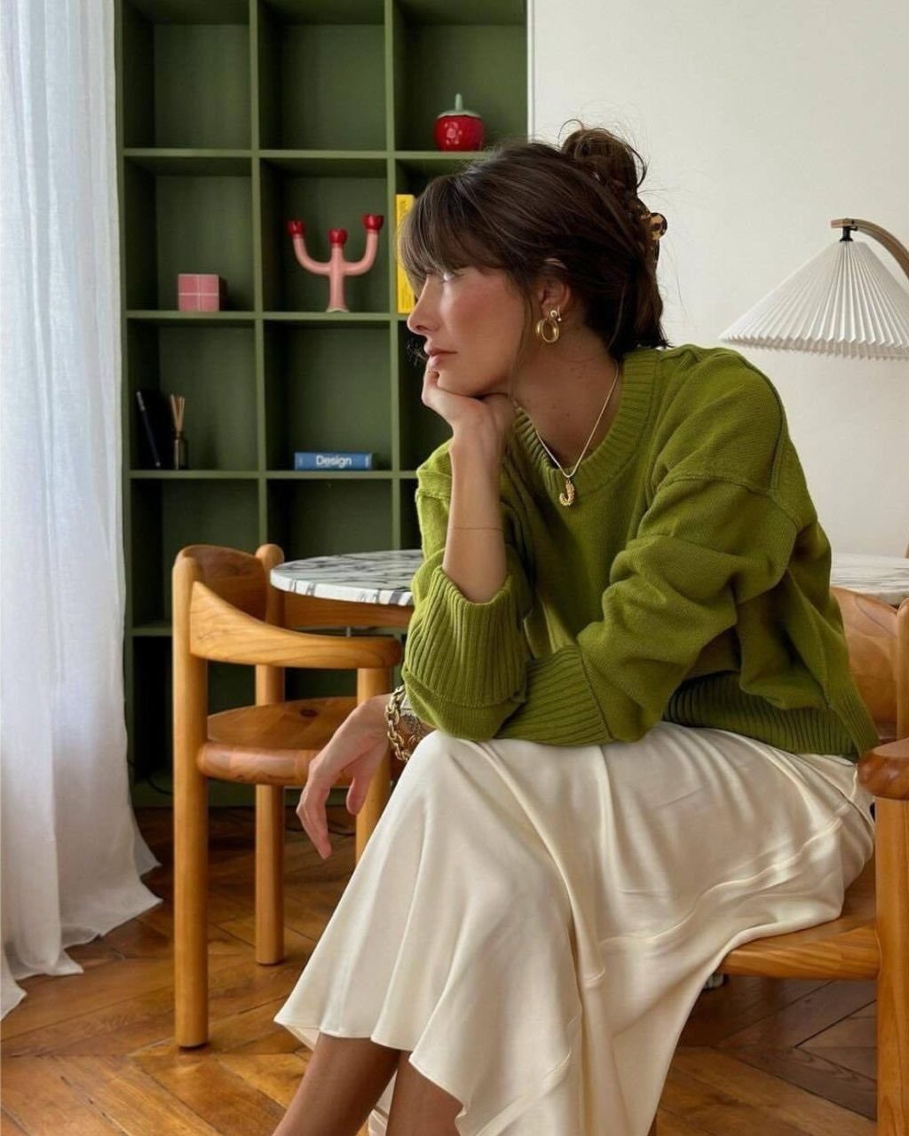 Woman wearing a moss green sweater and cream satin skirt sitting in a wooden chair.