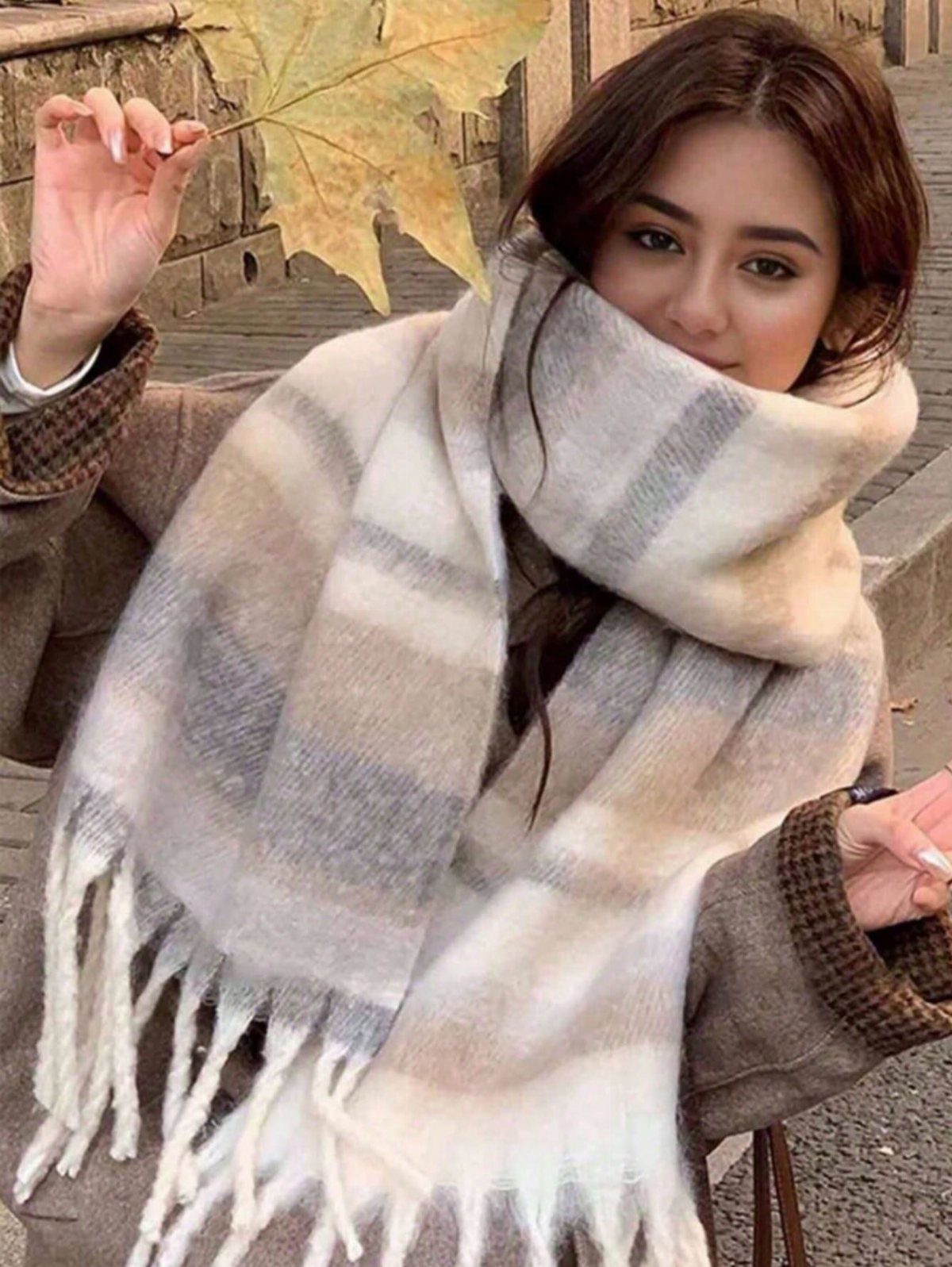 Woman wearing a large plaid scarf in neutral tones while holding a yellow autumn leaf.