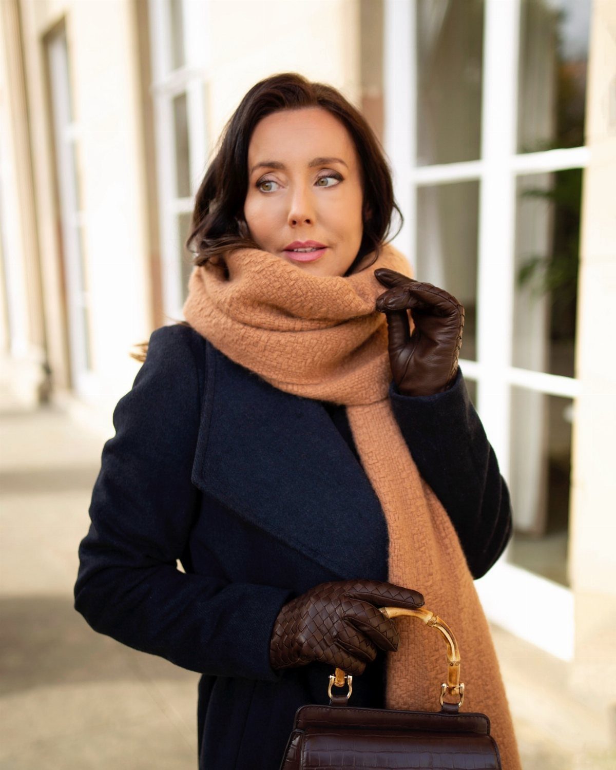 A woman wearing a navy coat, peach scarf, and brown leather gloves while holding a handbag.