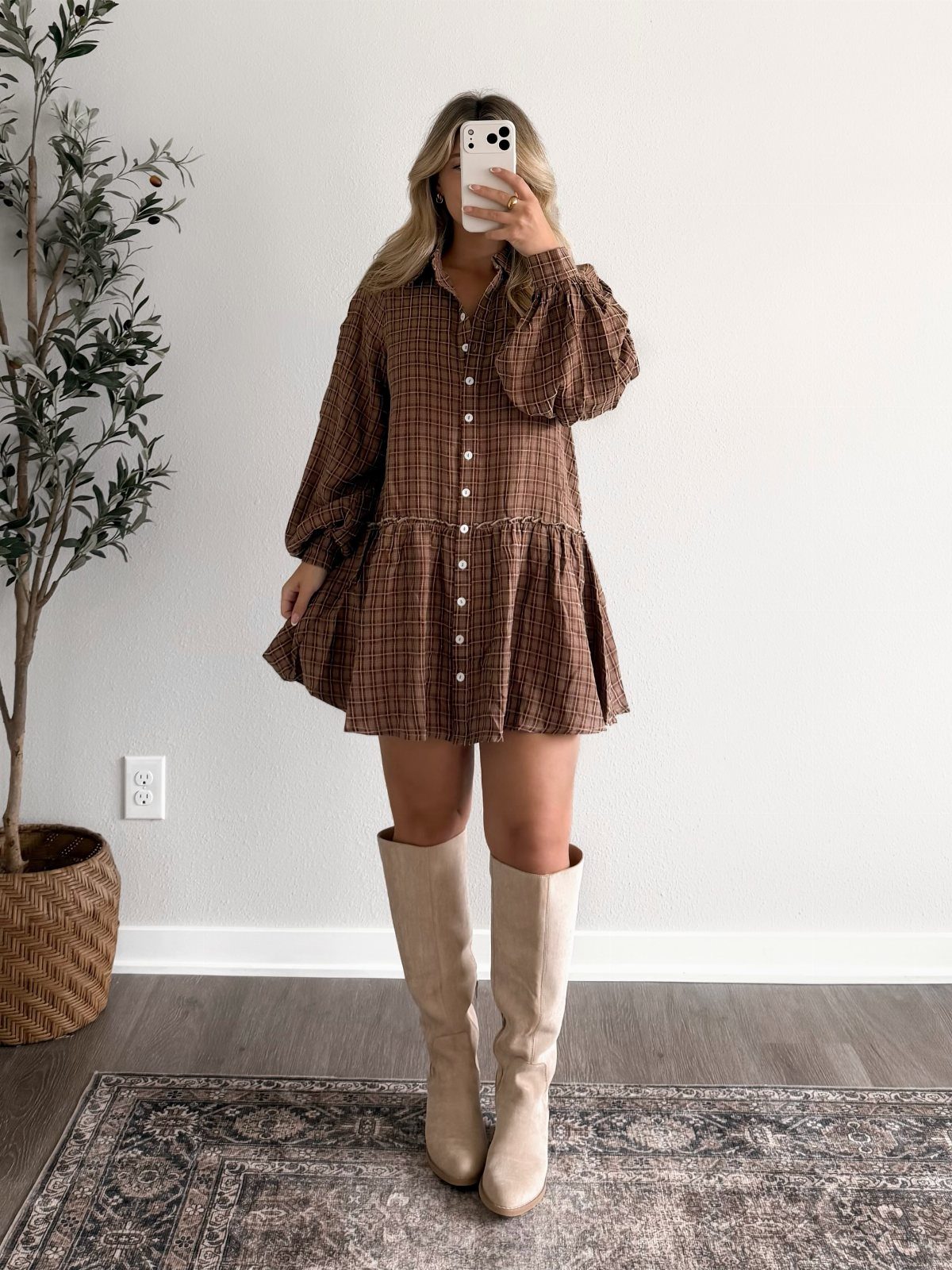Woman in a brown patterned mini dress and beige tall boots taking a mirror selfie.