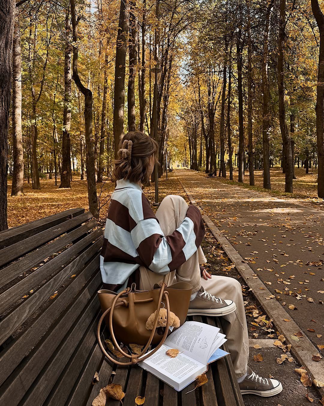 A person in autumn clothing sits on a park bench with a tan leather handbag.