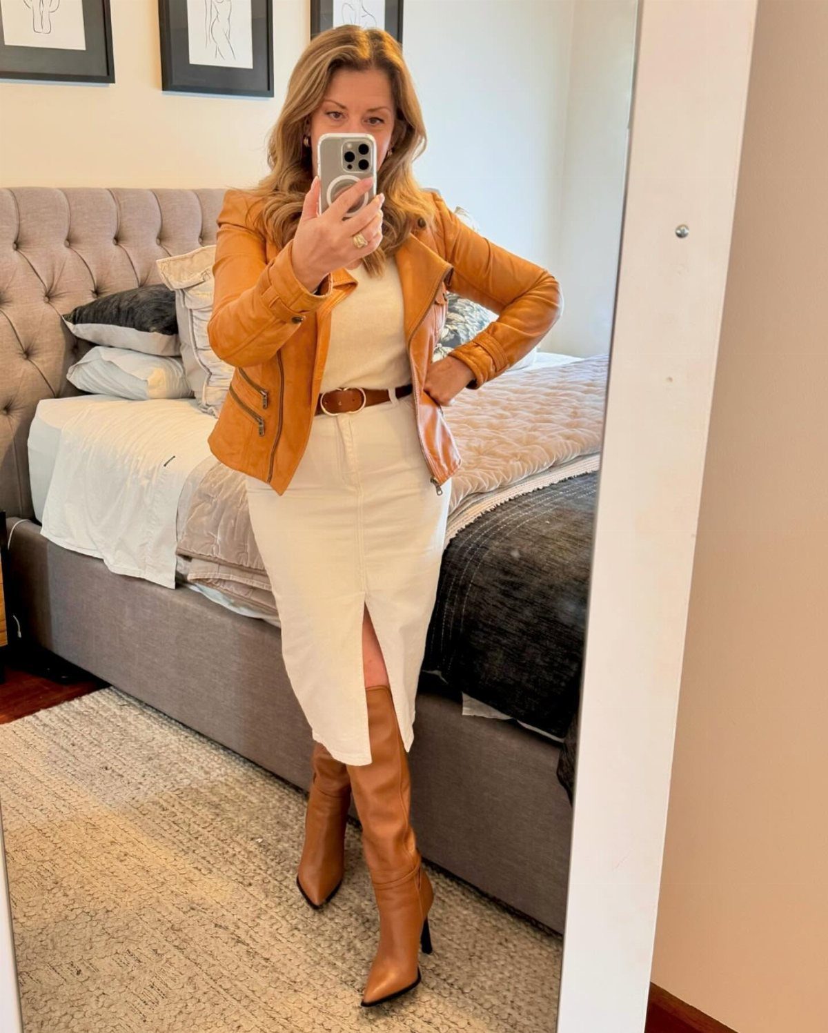 Woman in a tan leather jacket, cream skirt, and tall brown leather boots posing in a mirror.