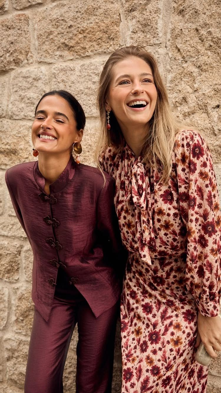 Two smiling women wearing warm-toned burgundy and patterned autumn clothing against a stone wall.