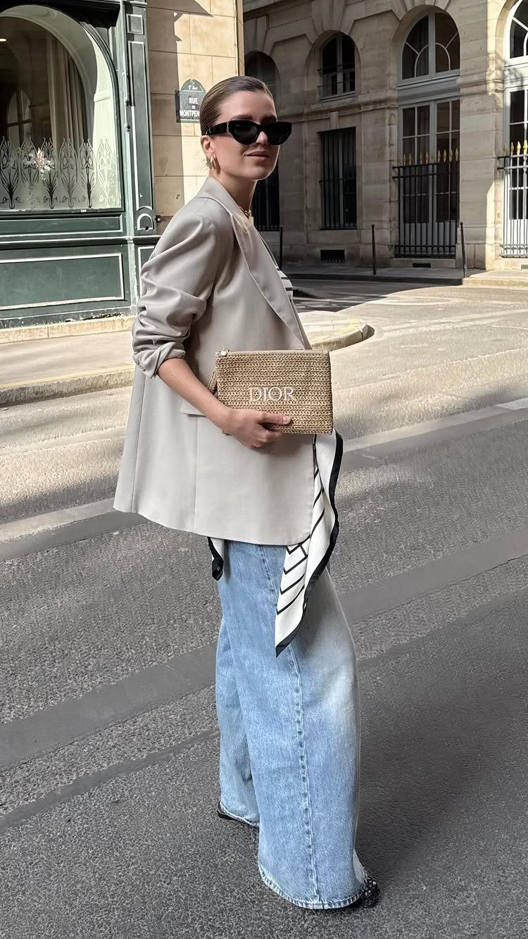 Woman in a beige blazer and wide-leg light blue denim jeans holding a straw clutch.