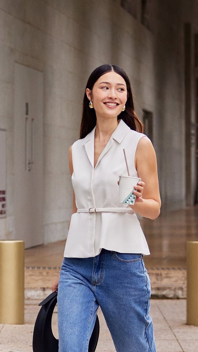 A smiling woman wearing a white sleeveless tailored vest and blue denim jeans walking outdoors.