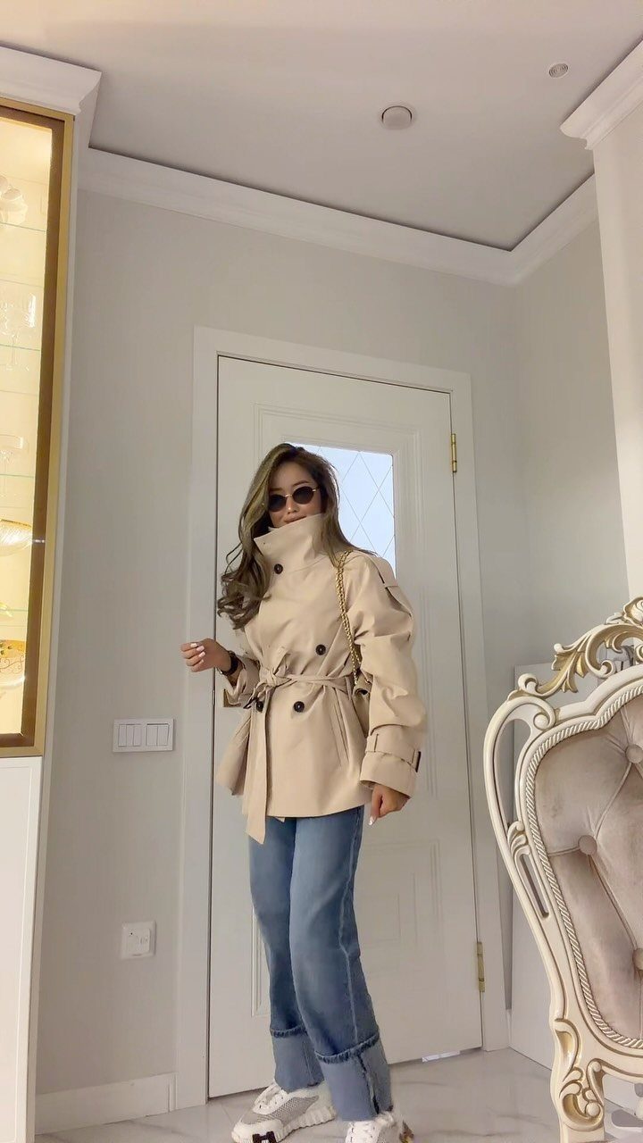 A woman wearing a beige belted trench coat and blue jeans standing indoors.