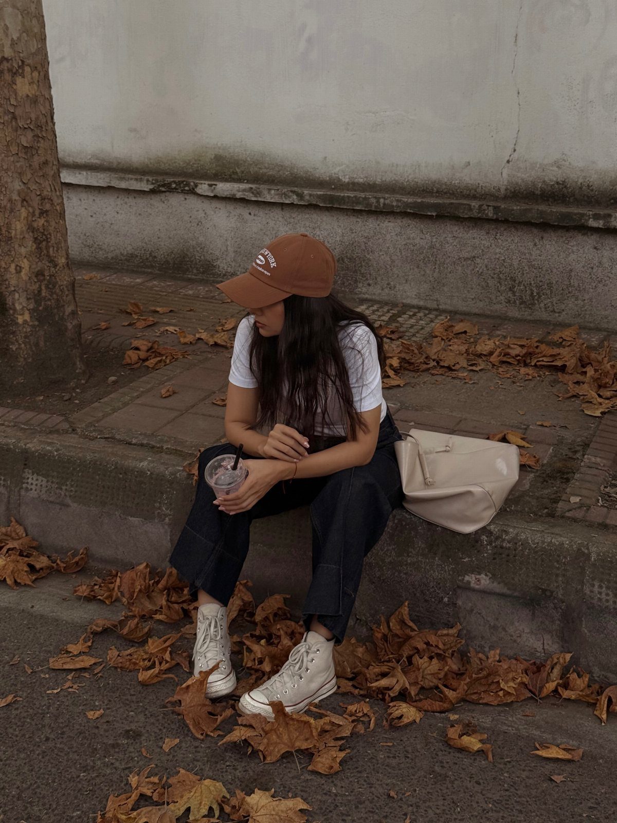 A person wearing a brown baseball cap and white sneakers sitting among autumn leaves.