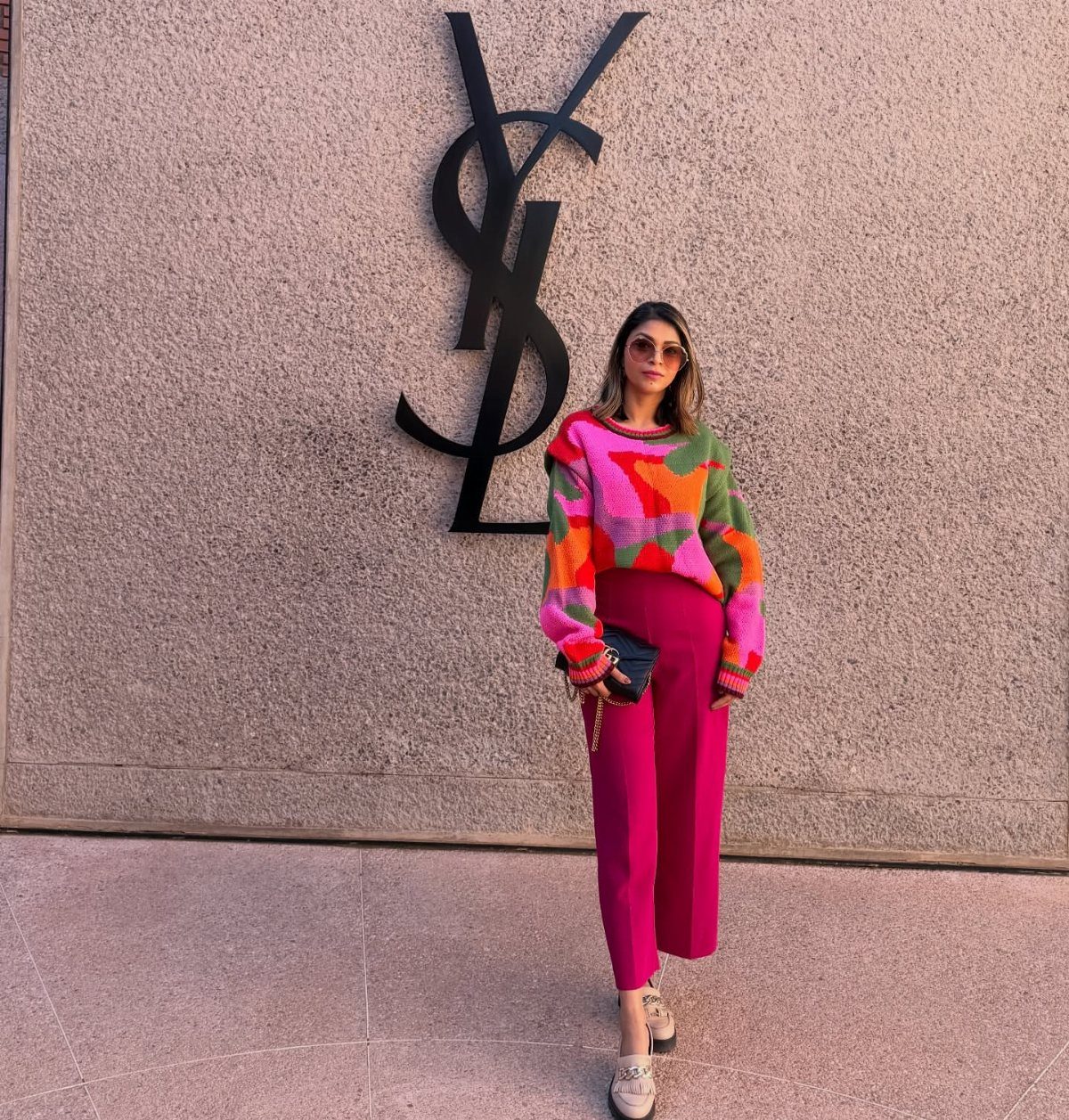 Woman wearing a colorful patterned sweater and bright magenta trousers standing before a large YSL logo.