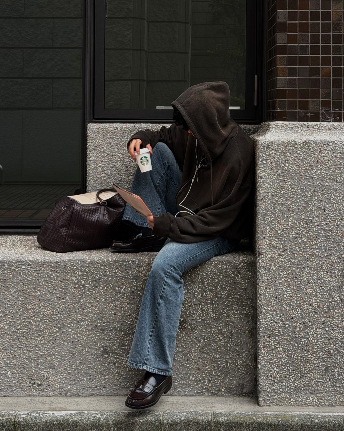 A person in a brown hoodie and blue jeans sitting on a concrete ledge holding coffee.