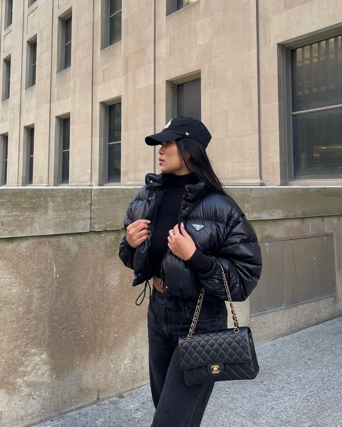 Woman in black puffer jacket, turtleneck, and denim jeans standing by a stone building.