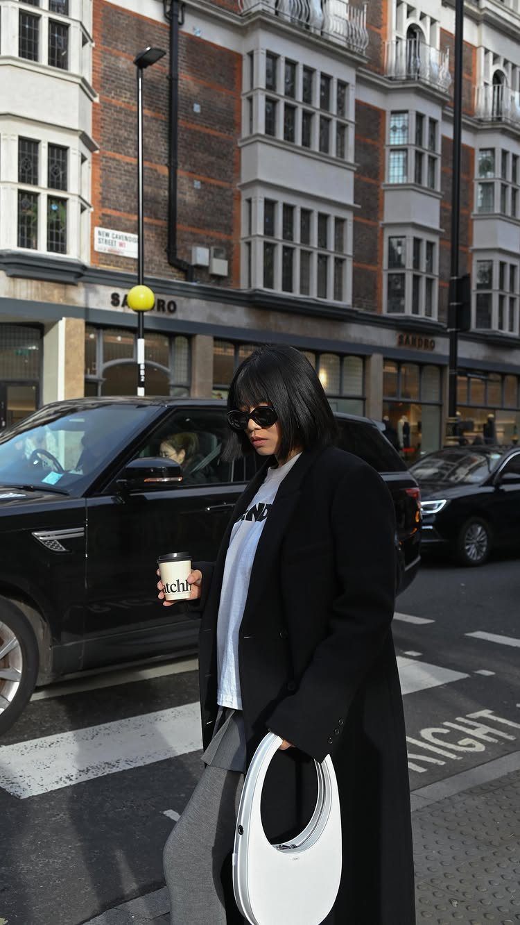 Woman in a long black chic coat walking through a city street with a white handbag.