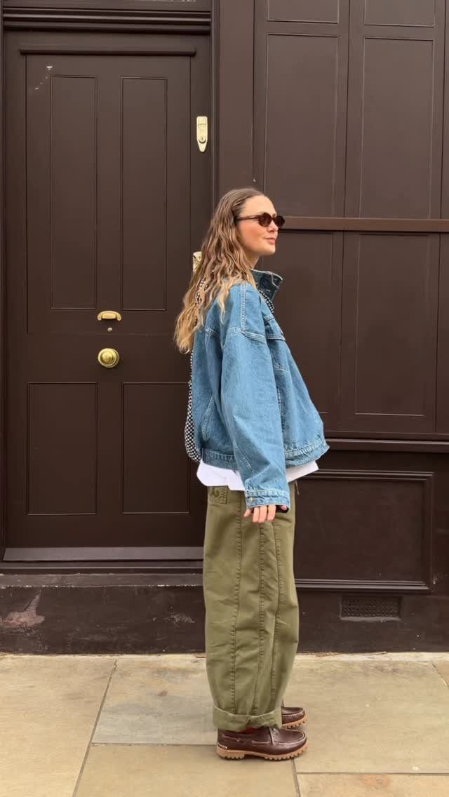 A woman wearing a denim jacket and olive trousers standing against a dark brown door.